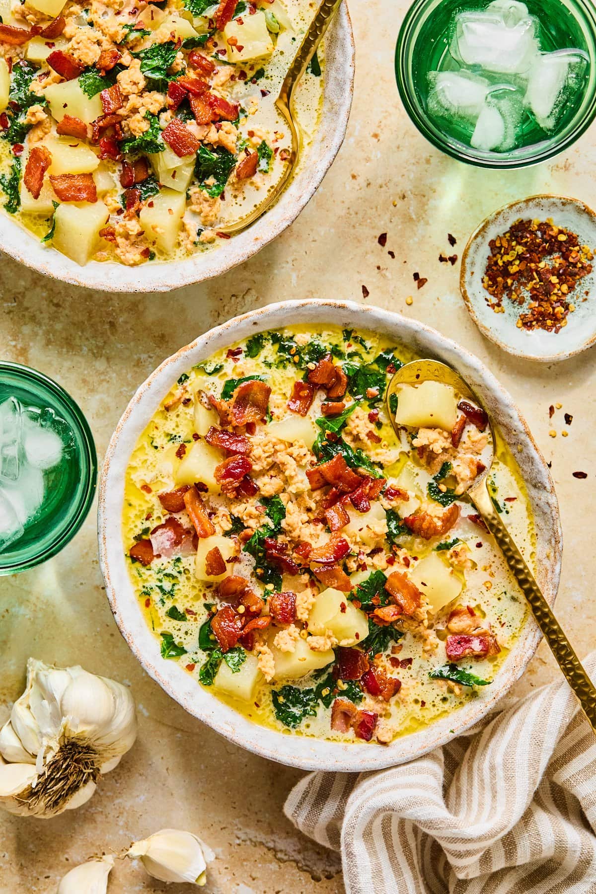 Two bowls of Instant Pot zuppa toscana with a spoon resting in the main bowl, surrounded by a garlic clove, a small bowl of red pepper flakes, and a tan and white striped dish towel.