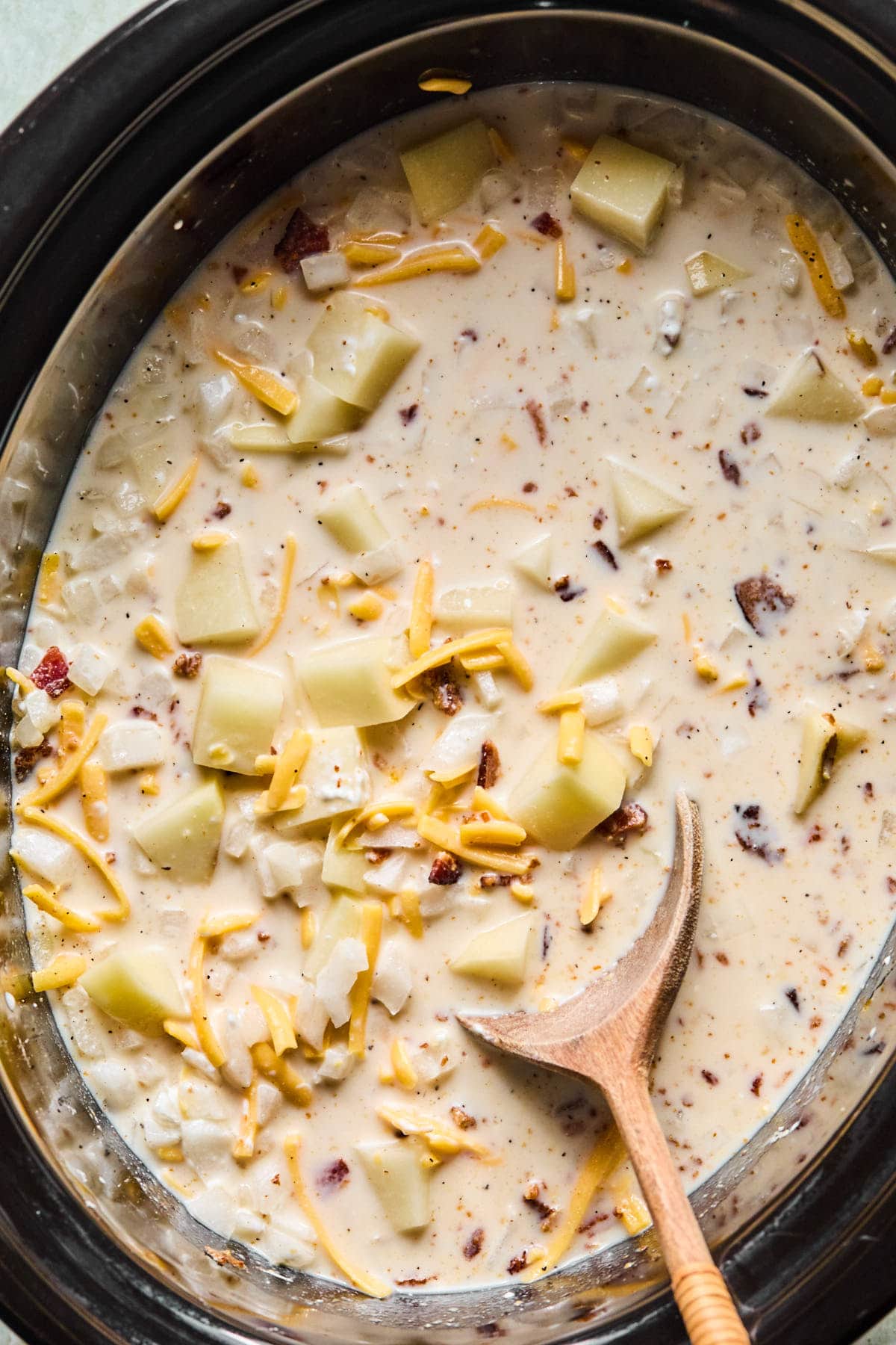 Cubed potatoes, chopped onion, garlic, bacon, and chicken broth in a slow cooker being stirred together.