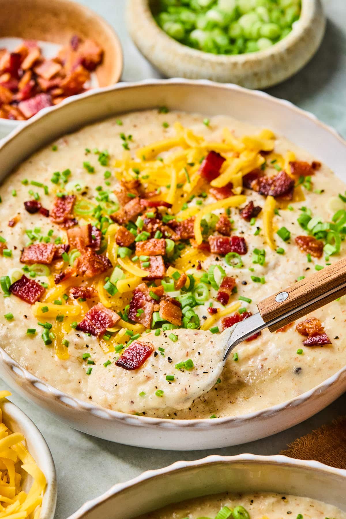 Bowl of crockpot potato soup topped with shredded cheddar, bacon, and sliced green onions, with a spoon lifting a bite.