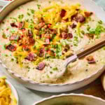 A bowl of crockpot potato soup garnished with cheddar, bacon, and green onions, with a spoon resting in it.