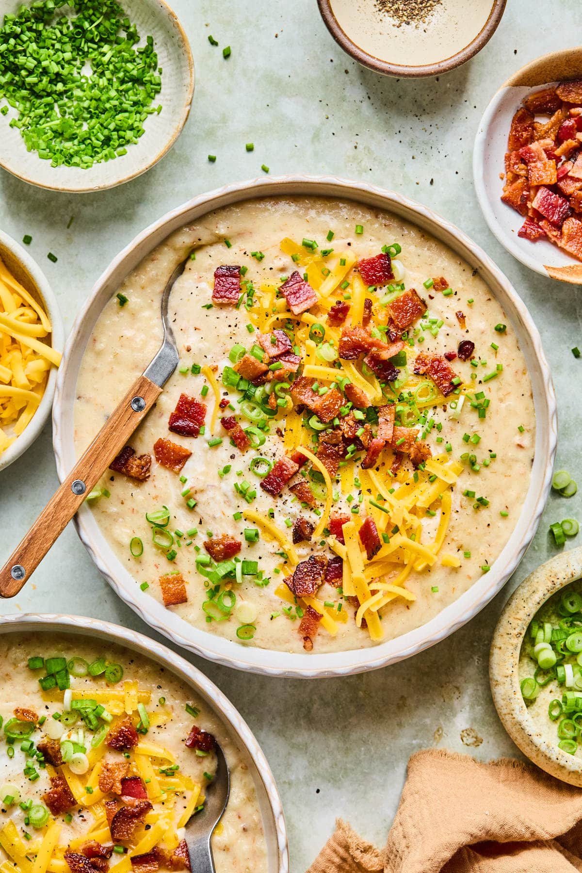 Two bowls of crockpot potato soup garnished with cheddar, bacon, and green onions, with a spoon resting in one bowl.