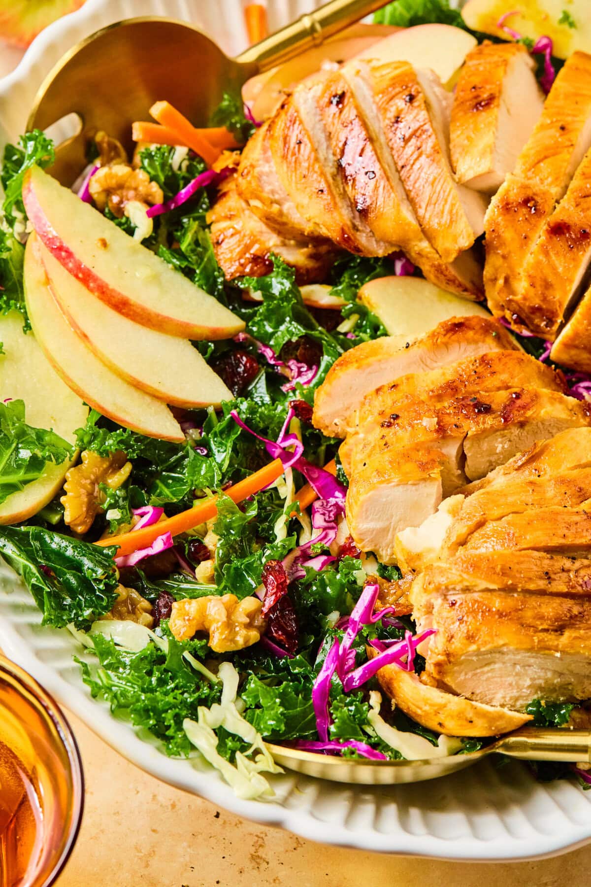 Close-up of winter kale salad showing sliced apples, shredded carrots, grilled chicken, and toasted walnuts.