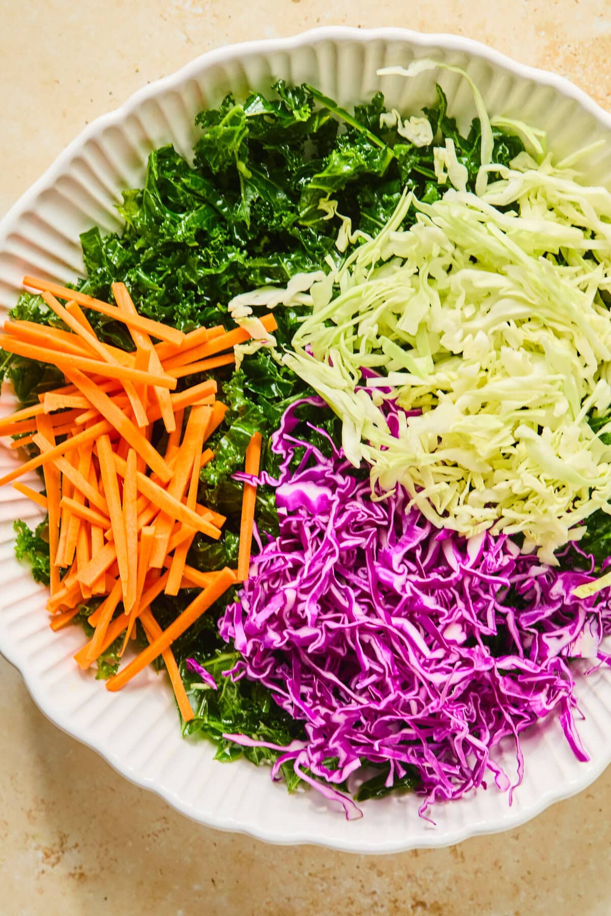 Shredded green cabbage, purple cabbage, and carrots added to a bowl of massaged kale.