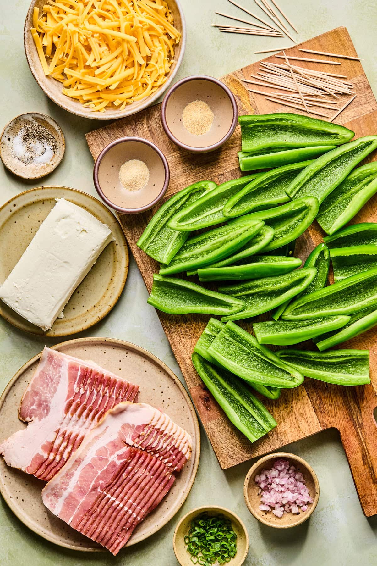 Ingredients for bacon wrapped jalapeno poppers arranged on a wooden cutting board with small plates and bowls.