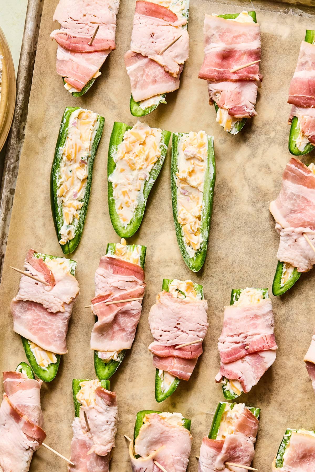 Fresh jalapeño halves filled with a creamy cheese mixture, smoothed level on top, with some wrapped in bacon and secured with toothpicks, arranged on a parchment-lined baking sheet.