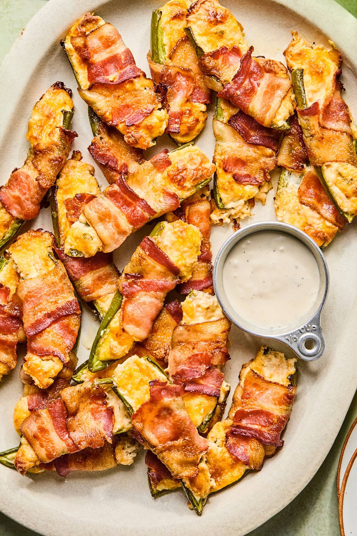 Bacon wrapped jalapeno poppers served on a white platter with a silver cup of homemade ranch dipping sauce.