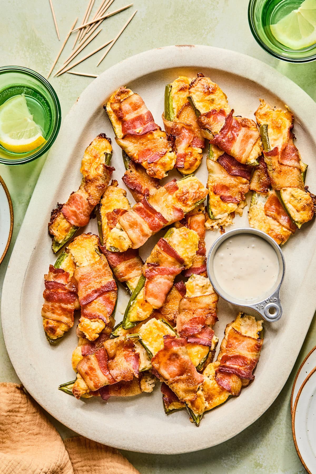 Bacon wrapped jalapeno poppers served on a white platter with a silver cup of homemade ranch dipping sauce.
