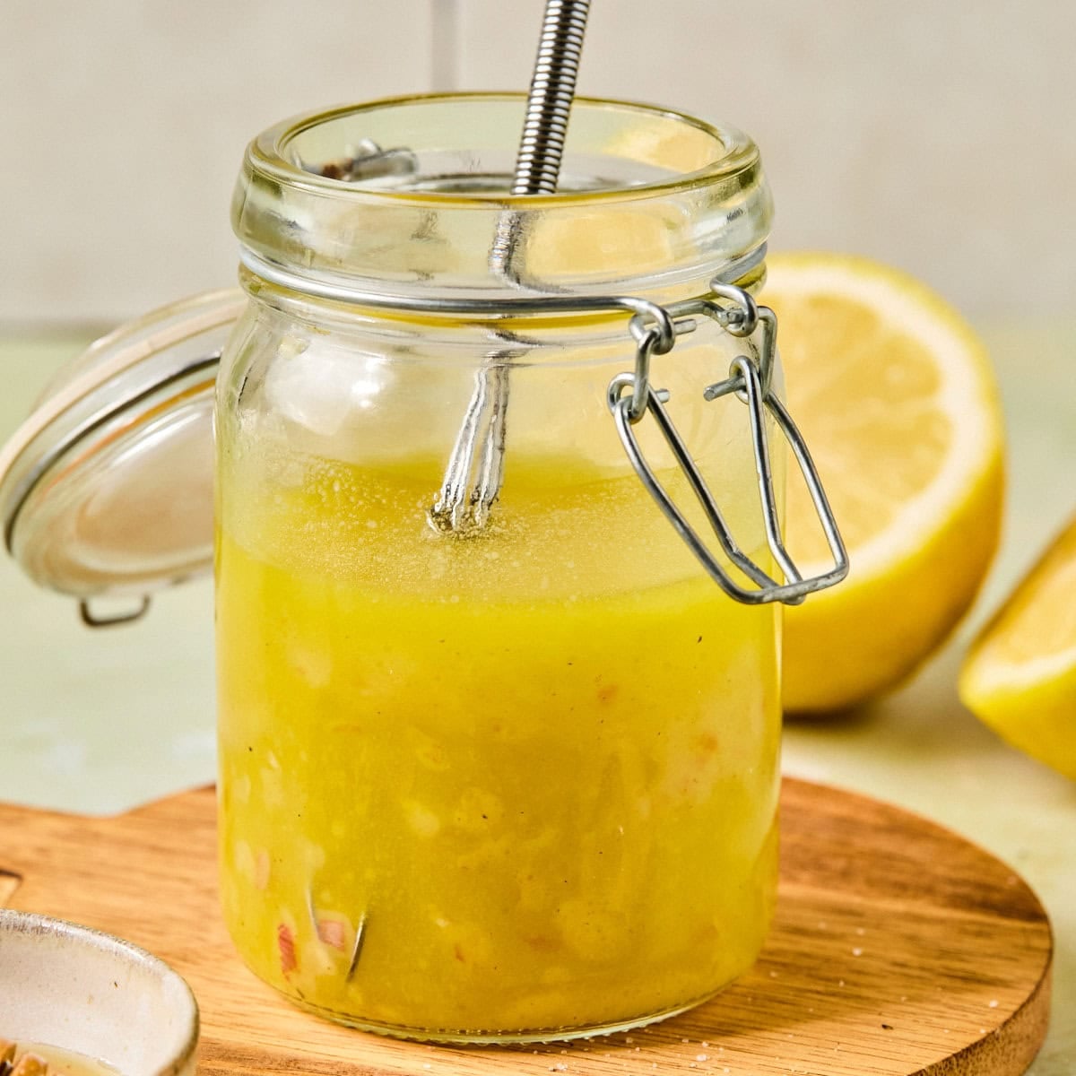 Mason jar of homemade lemon vinaigrette with a whisk, set on a round wooden board surrounded by fresh lemons.