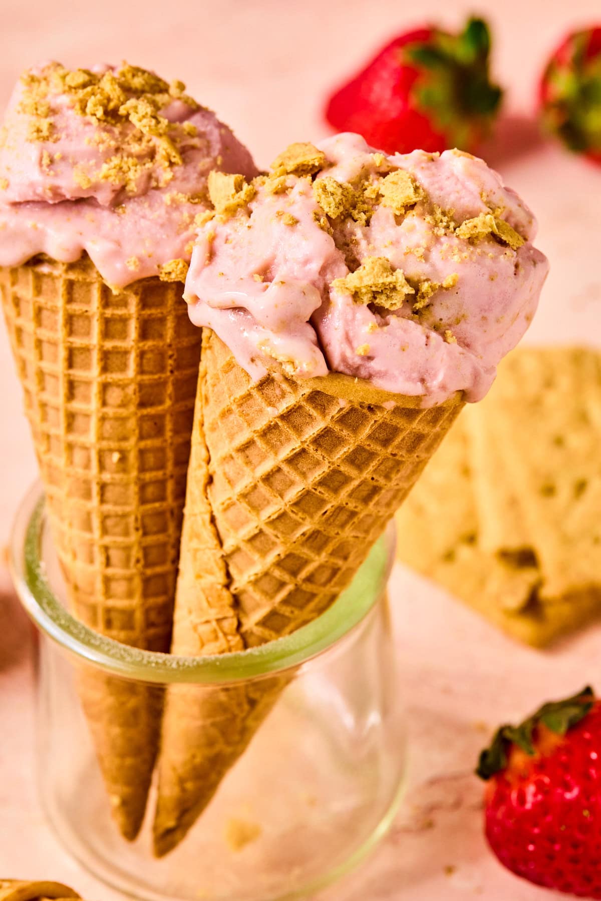 Two waffle cones filled with Ninja Creami strawberry ice cream, topped with graham cracker crumbs, set against a soft pink background.