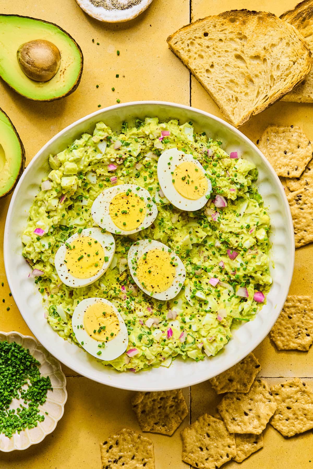 Avocado egg salad in a serving bowl topped with halved hard-boiled eggs, surrounded by crackers, toast, sliced avocado, and a nesting bowl of chopped chives.