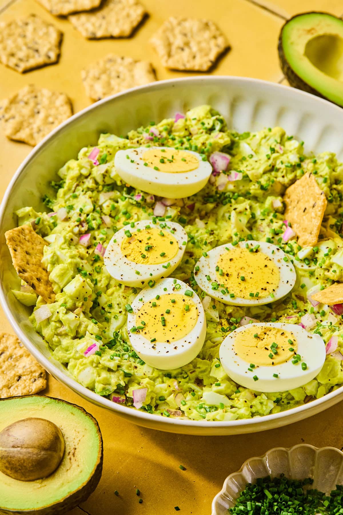 Avocado egg salad in a bowl topped with halved eggs, served with crackers, half an avocado, and a small bowl of fresh chives.