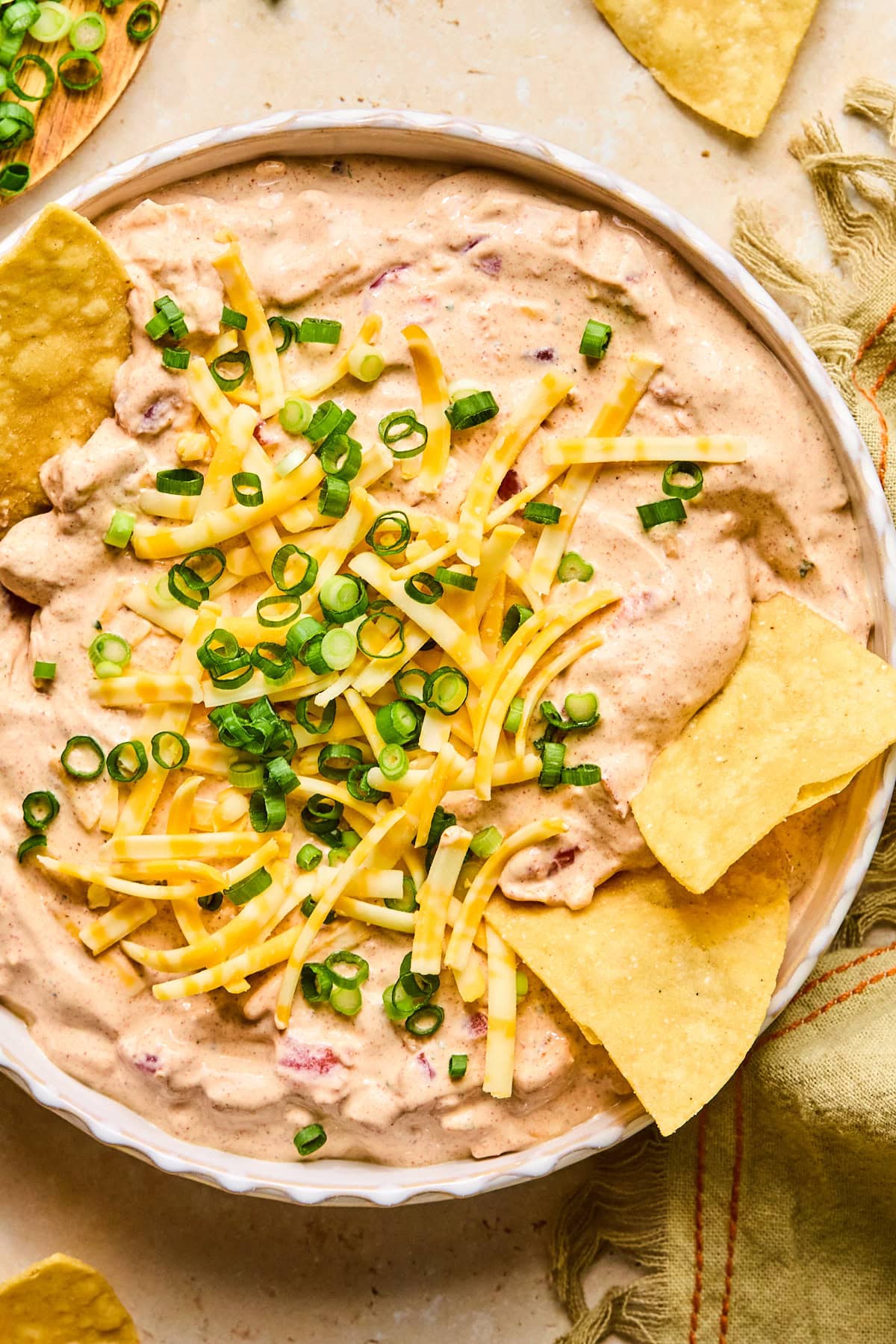 Boat dip topped with shredded cheese and green onions, with tortilla chips in the dip and scattered behind the bowl.