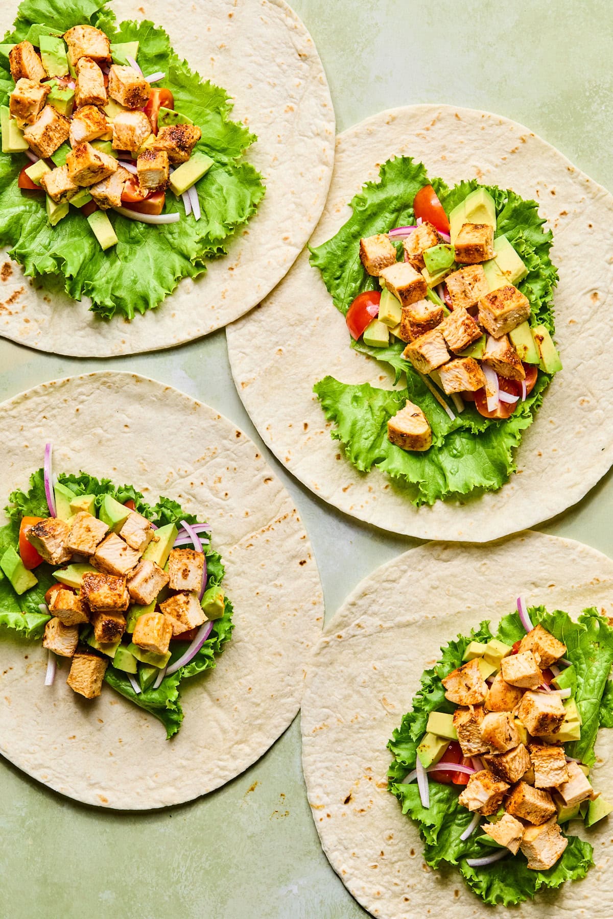 Four tortillas laid out on a countertop, each topped with lettuce, tomatoes, red onion, avocado, and chopped chicken.