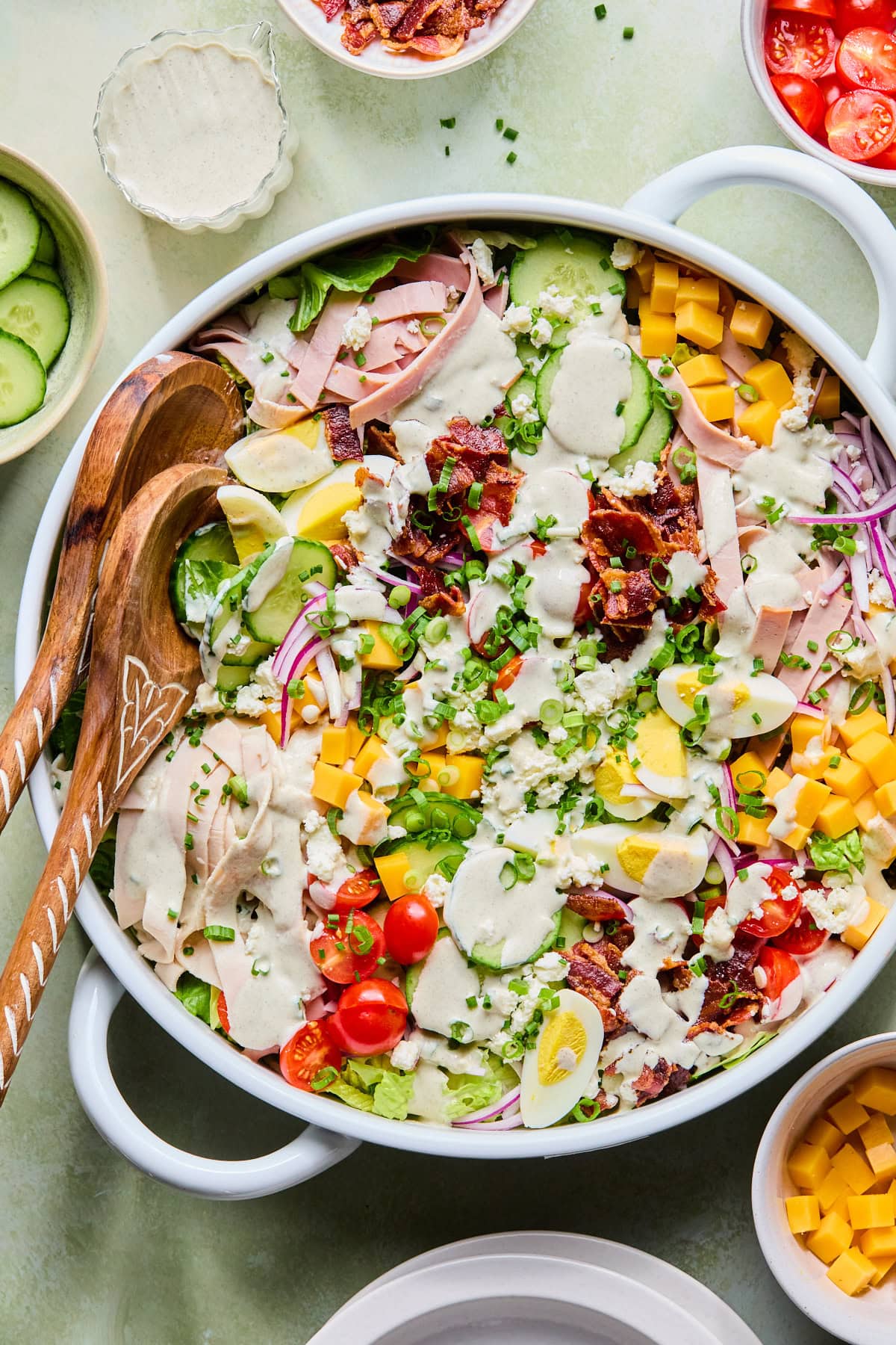 Chef’s salad in a large white serving bowl with creamy Italian dressing drizzled on top of it and serving spoons resting inside.
