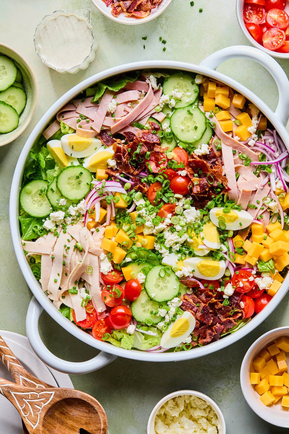 Chef’s salad in a large white serving bowl with romaine, iceberg lettuce, turkey, ham, bacon, cheddar cheese, feta, cucumbers, cherry tomatoes, and hard-boiled eggs. Small bowls of cucumbers, bacon, cherry tomatoes, and creamy Italian dressing sit in the background.