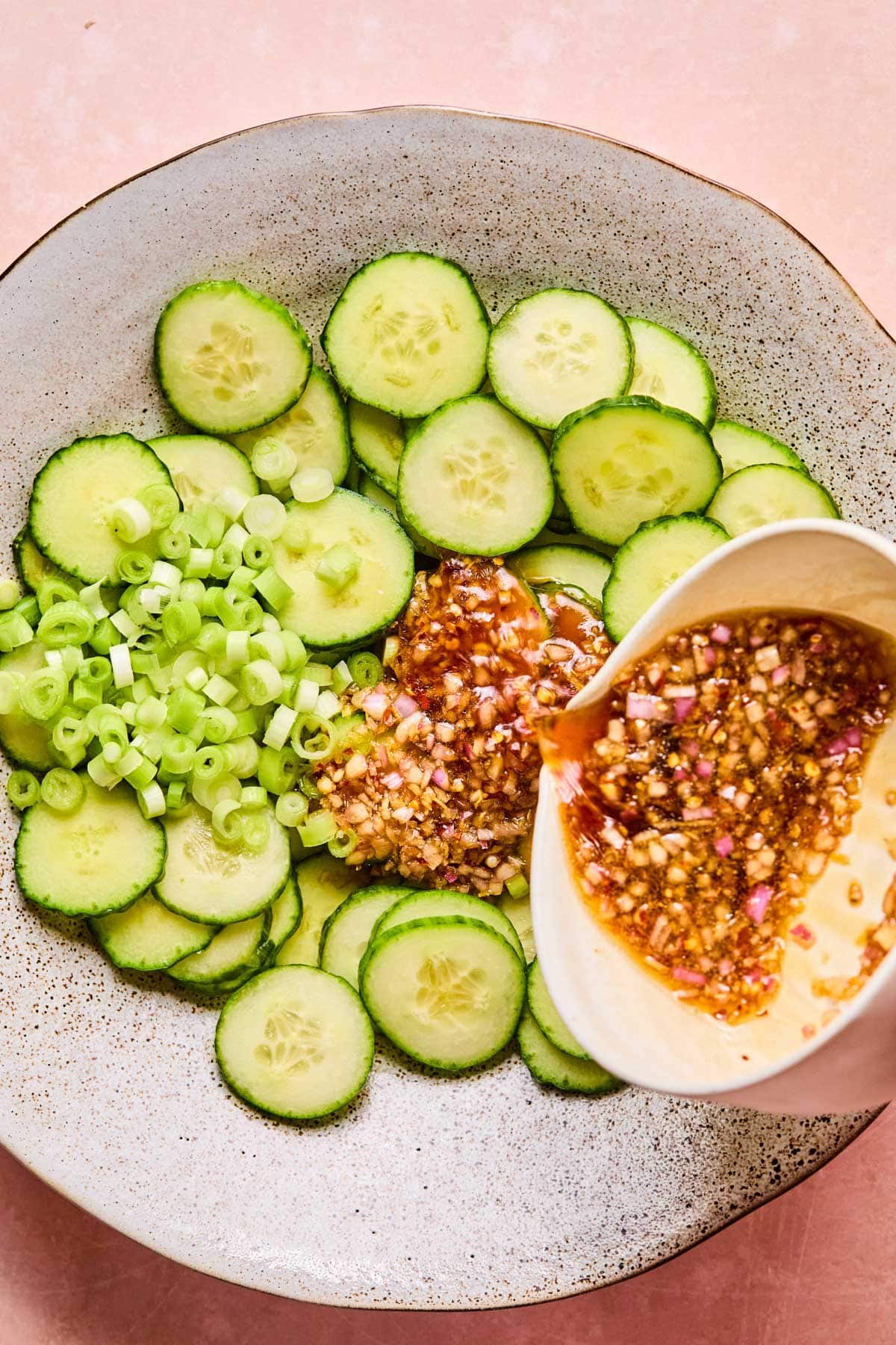 Dressing being poured over the thinly sliced mini cucumbers and chopped green onions.