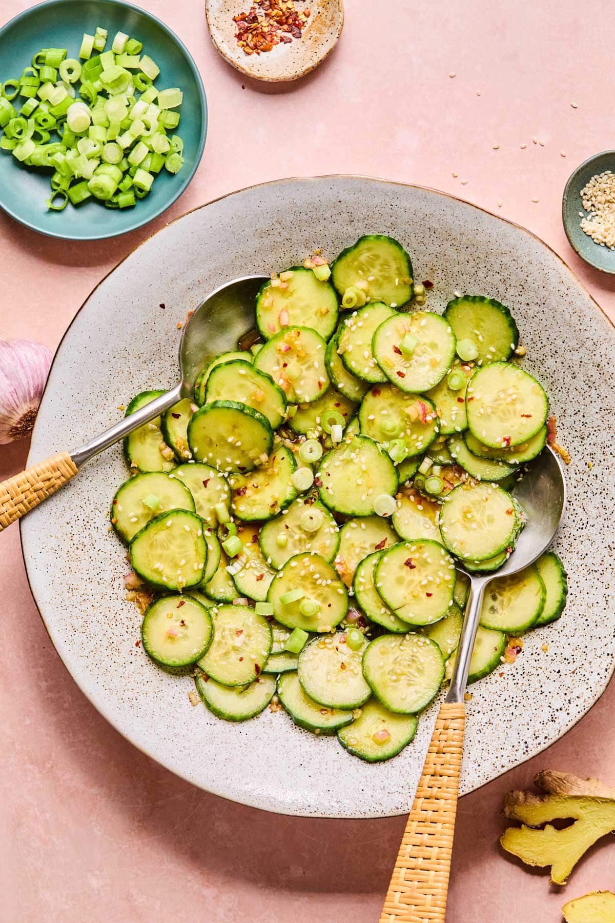 Bowl of Asian cucumber salad with serving spoons, garnished with green onions, sesame seeds, and red pepper flakes, with small bowls of toppings in the background.