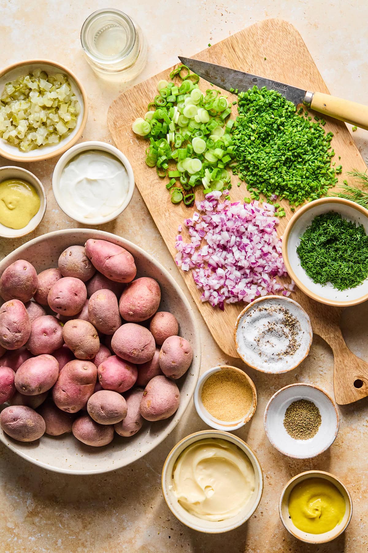 Ingredients for red potato salad recipe arranged on a countertop with red potatoes, dill pickles, fresh herbs, onions, and dressing ingredients. A knife rests beside chopped fresh ingredients on a wooden cutting board.