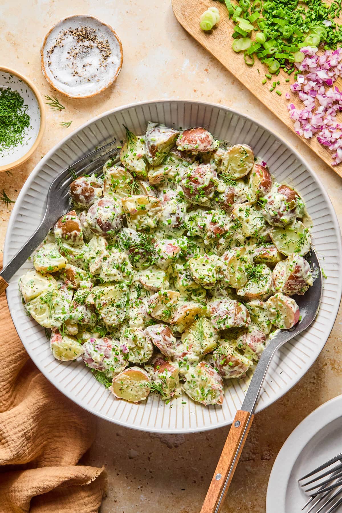 Bowl of skin-on red potato salad garnished with fresh dill and chives. A serving fork and knife sit in the salad on either side of the bowl.