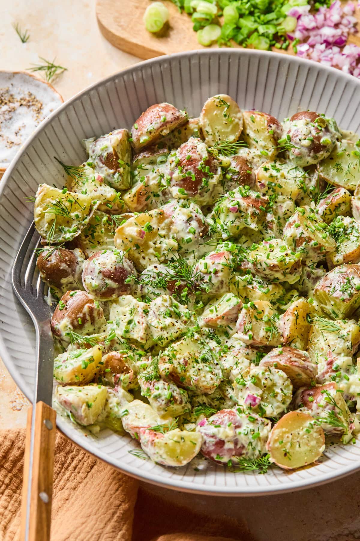 Close-up of creamy skin-on red potato salad garnished with fresh dill and chives, with a serving fork and knife placed inside of the bowl.