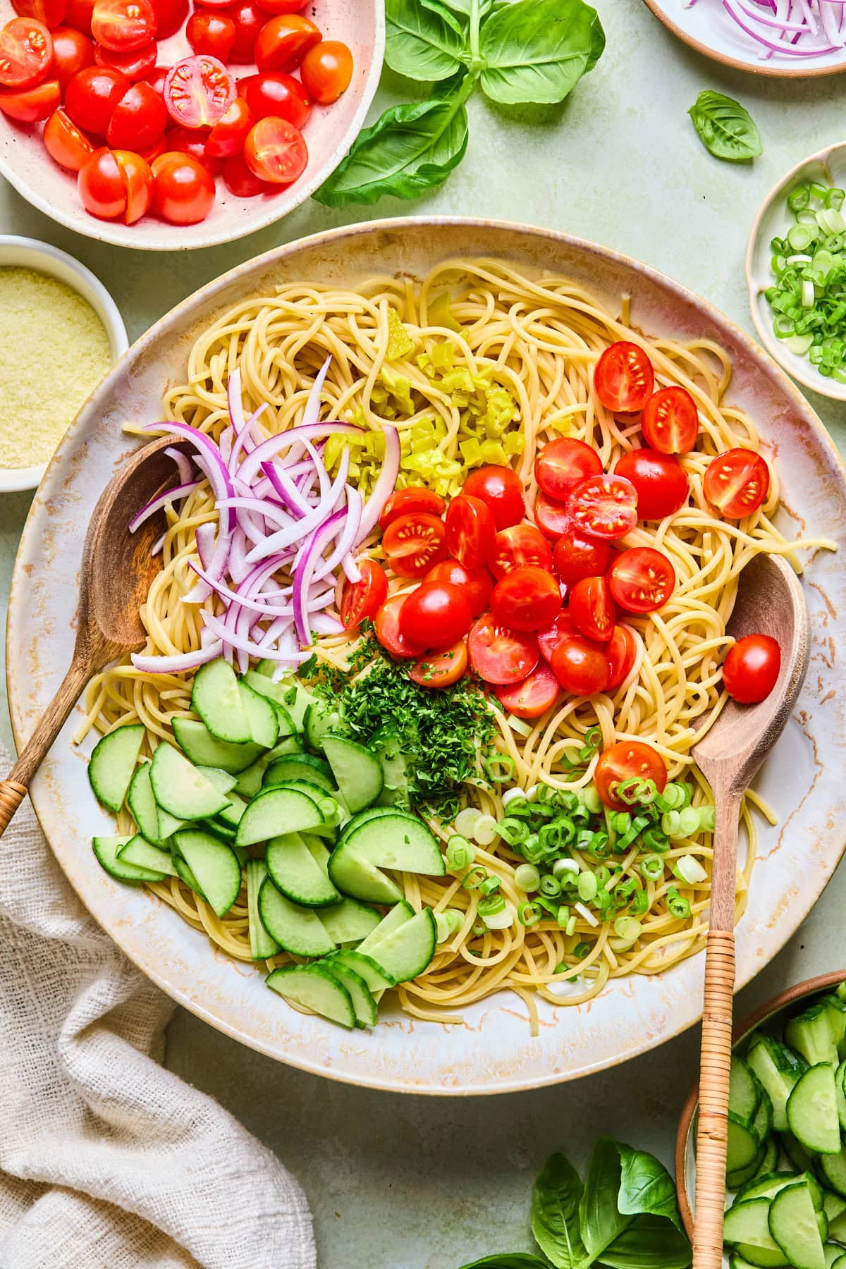 Cooked spaghetti ready to be tossed with grape tomatoes, cucumbers, red onion, green onions, pepperoncinis, and fresh parsley in a large mixing bowl.