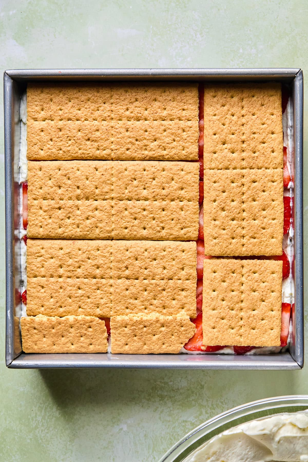 An 8x8-inch baking dish lined with a thin layer of cream filling and graham crackers arranged in a single layer for strawberry icebox cake.