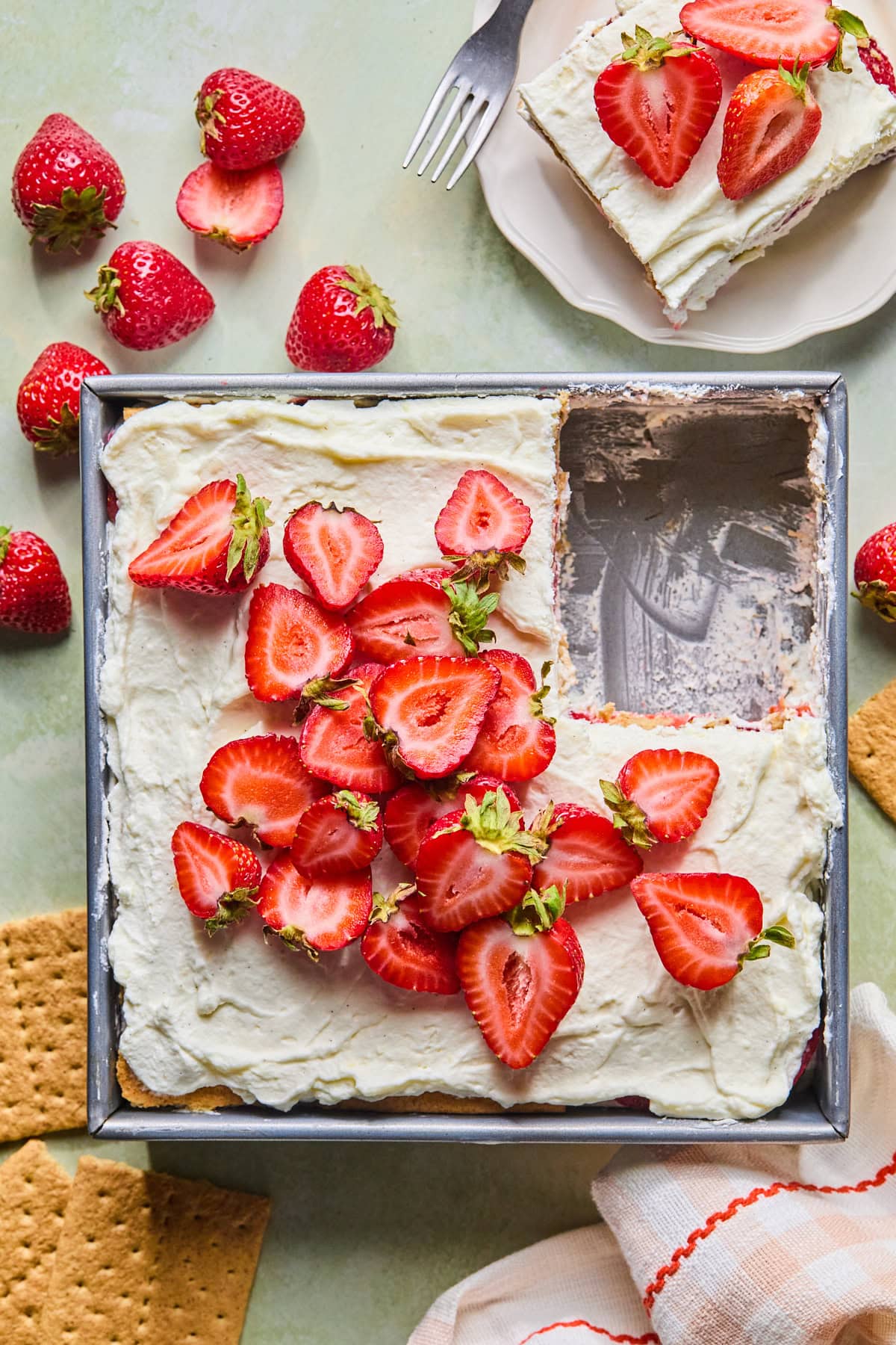 Strawberry icebox cake in an 8x8 pan with a corner piece removed, surrounded by fresh strawberries and graham crackers. A plated slice of strawberry icebox cake with a fork sits in the background.