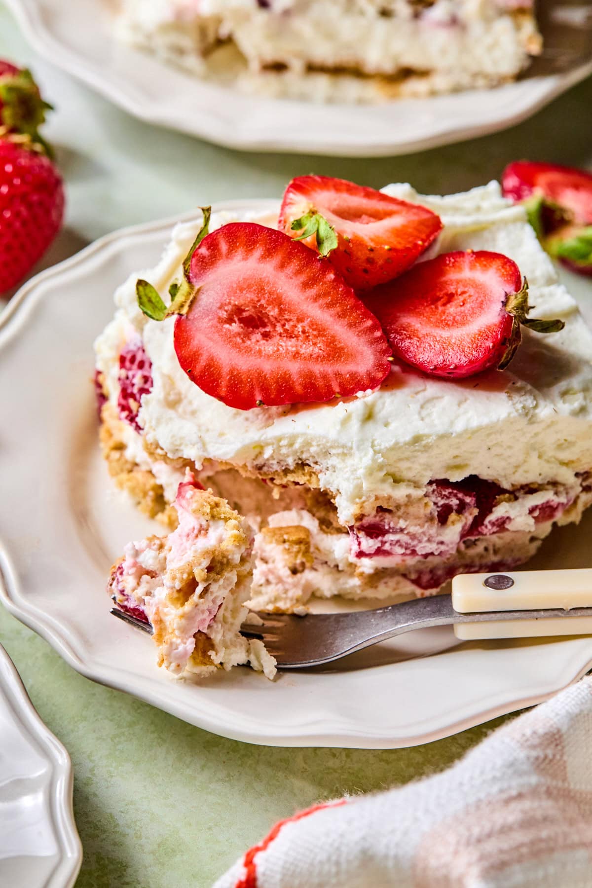 Close-up of a slice of strawberry icebox cake with cream cheese filling on a plate, topped with fresh strawberries and a fork holding a bite.