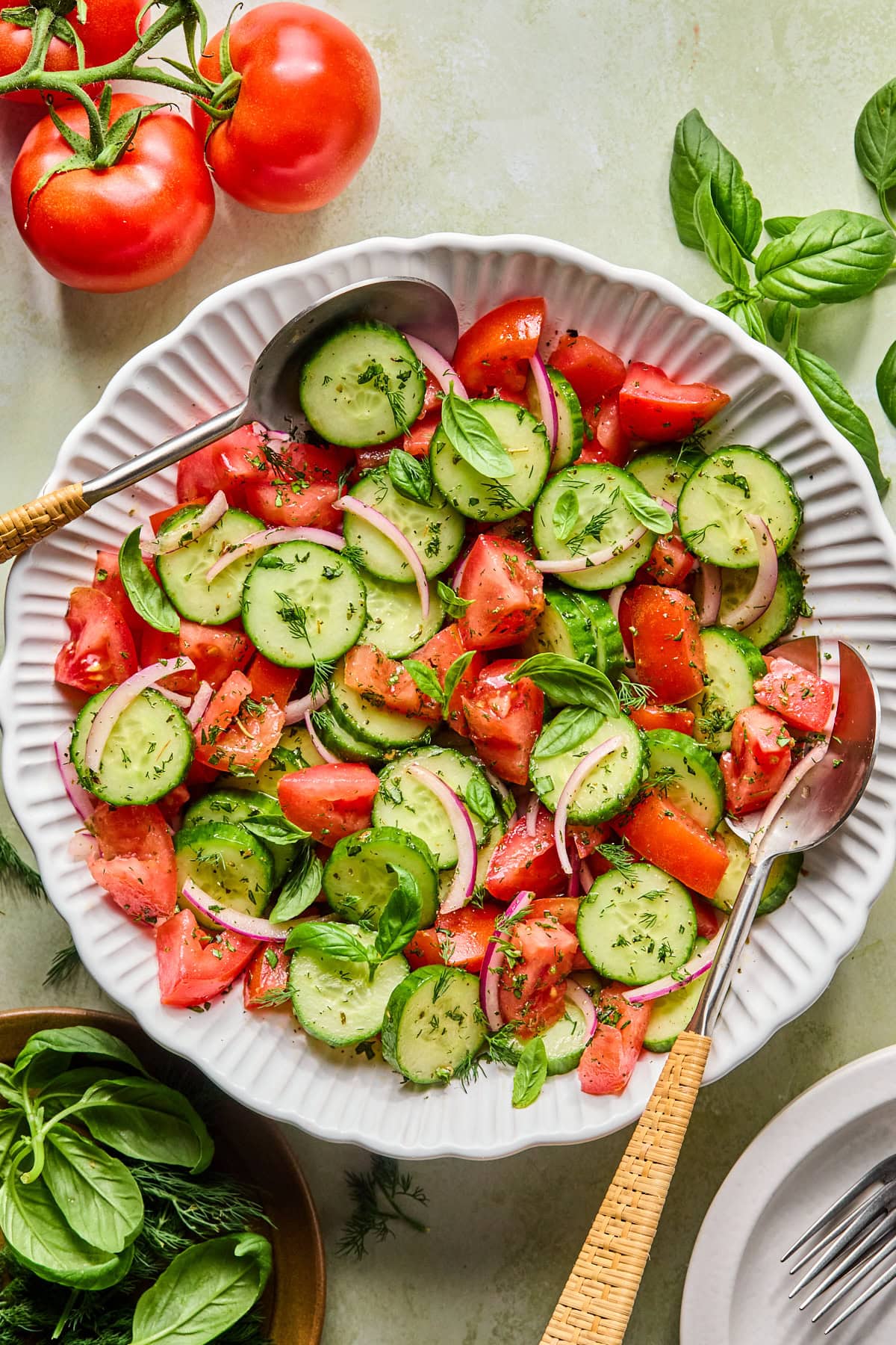 Tomato cucumber salad garnished with fresh basil and dill in a white serving bowl with serving spoons.