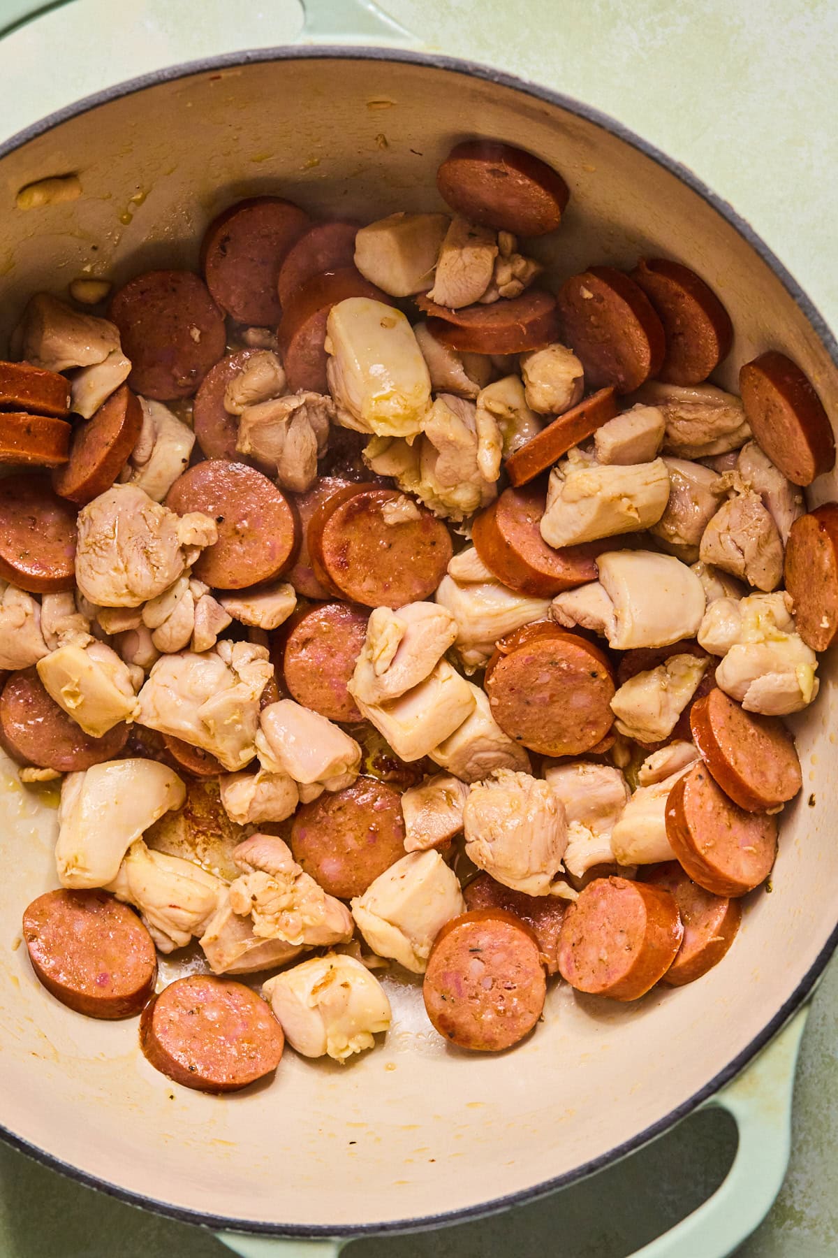 Chicken and sliced andouille sausage browning in a large skillet with oil.