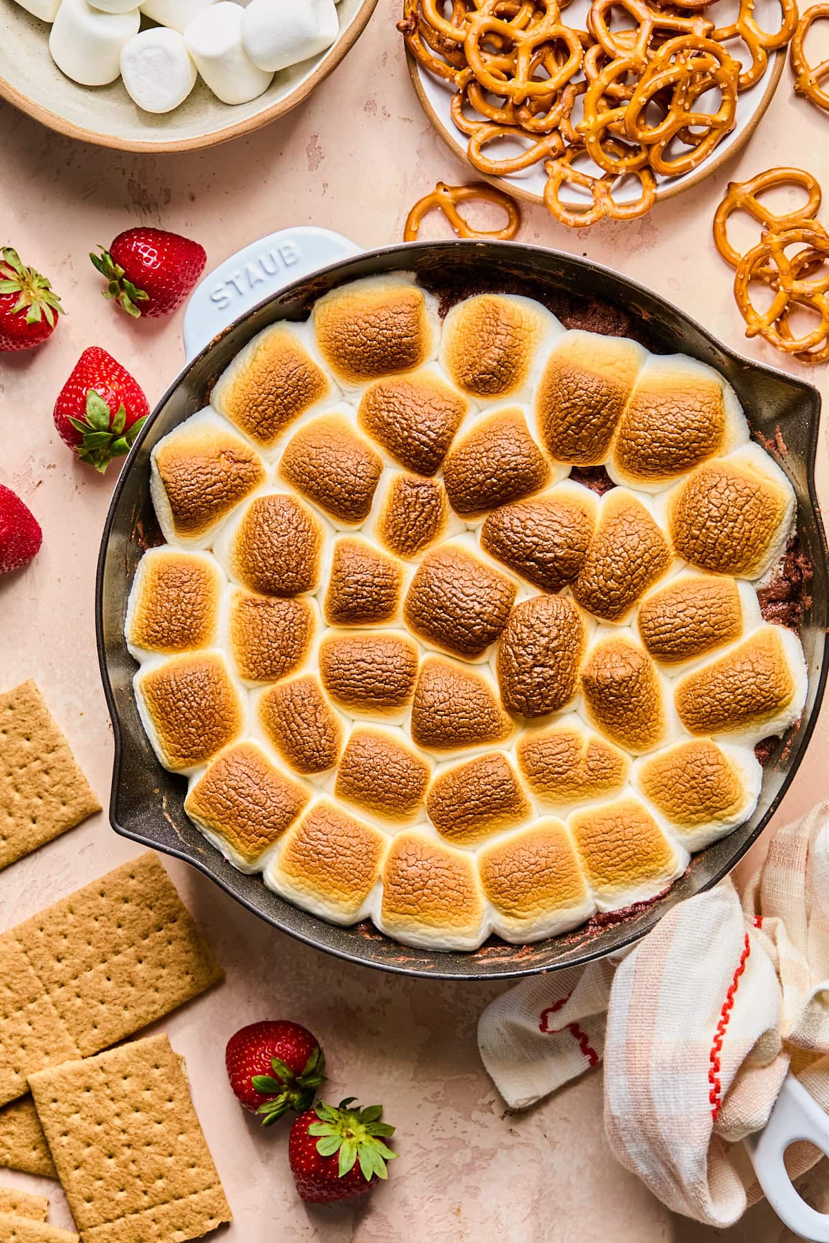 Overhead view of creamy s'mores dip in a cast iron skillet with golden toasted marshmallows, graham crackers, strawberries, pretzels, and marshmallows for dipping.