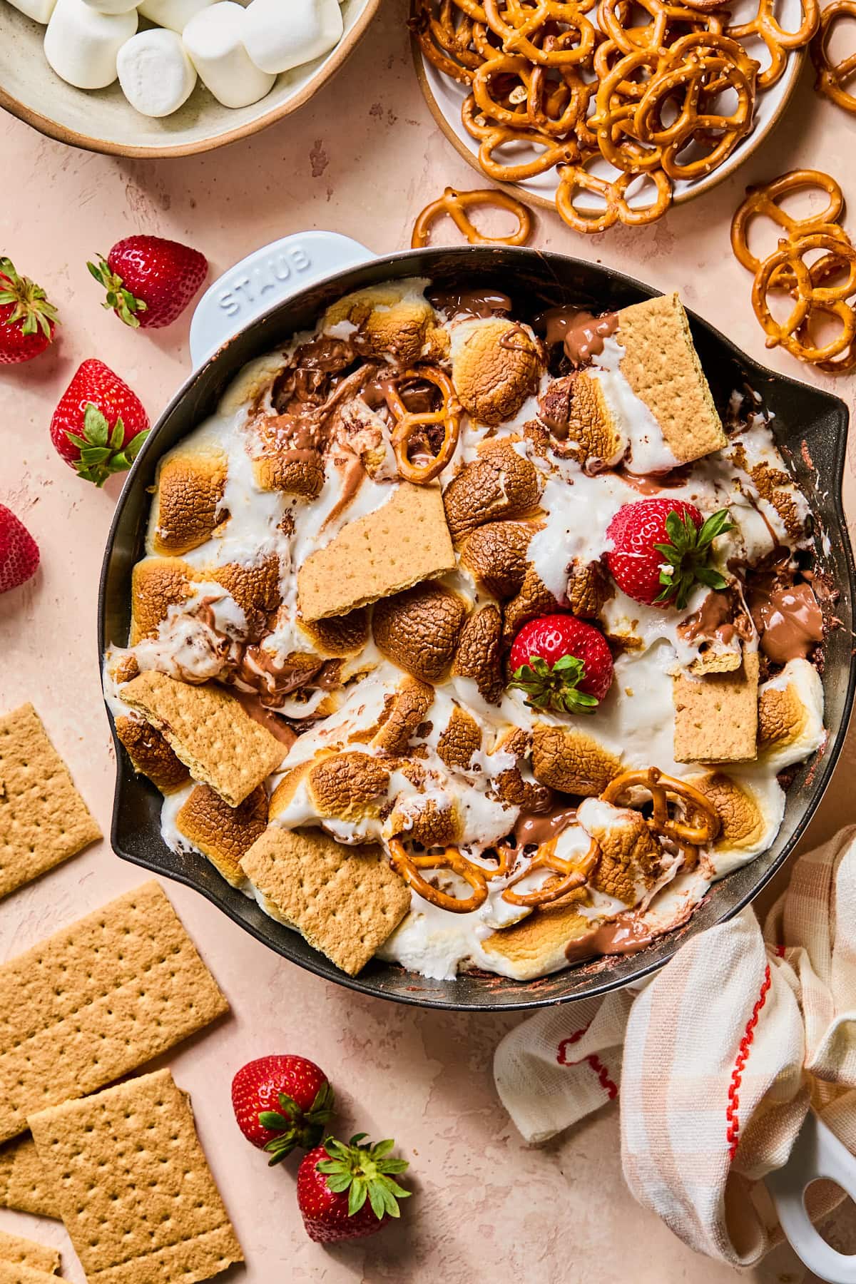 Overhead view of smores dip in a cast iron skillet with strawberries, graham crackers, and pretzels stuck in it.