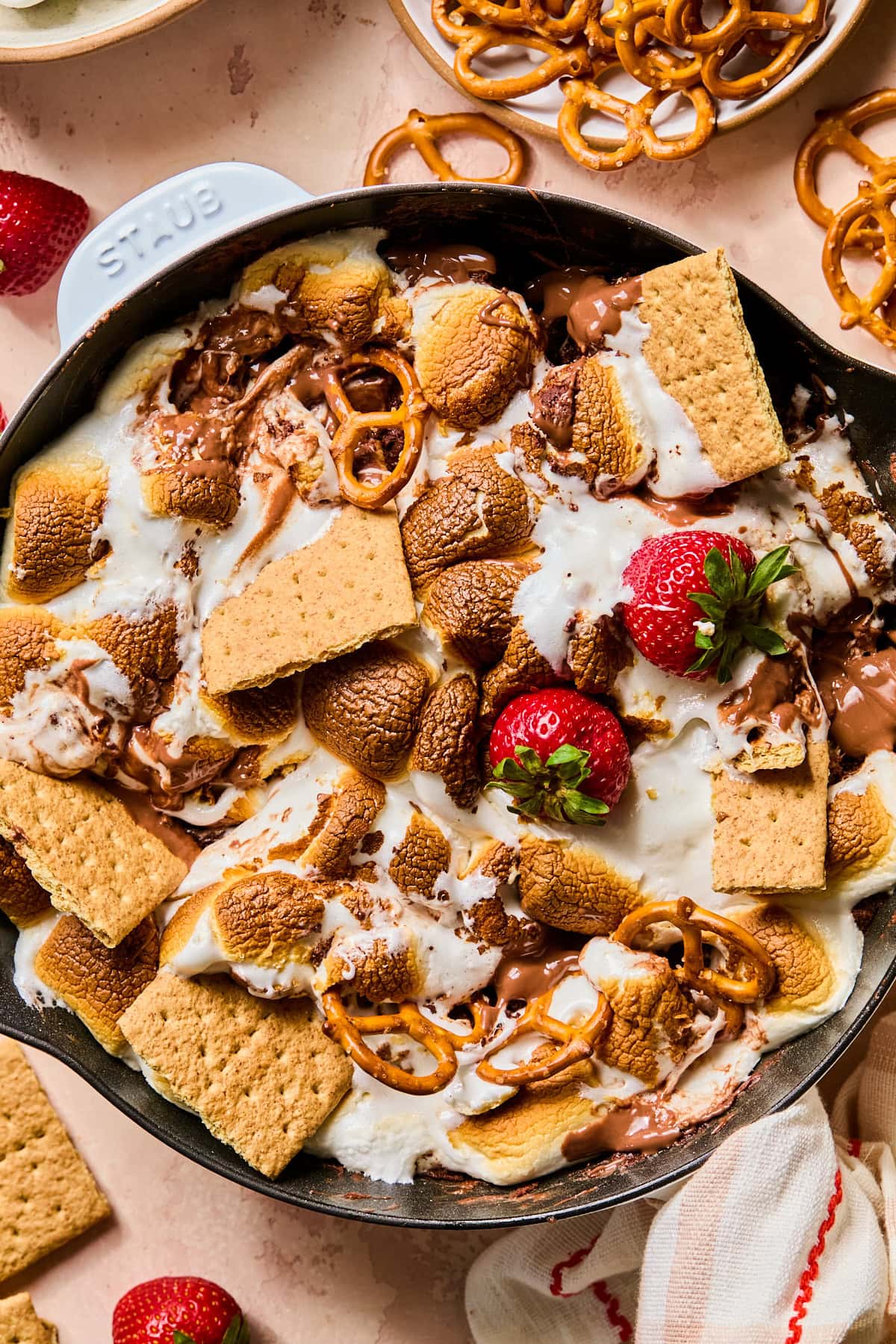 Overhead view of smores dip in a cast iron skillet with graham crackers, pretzels, and strawberries in it.