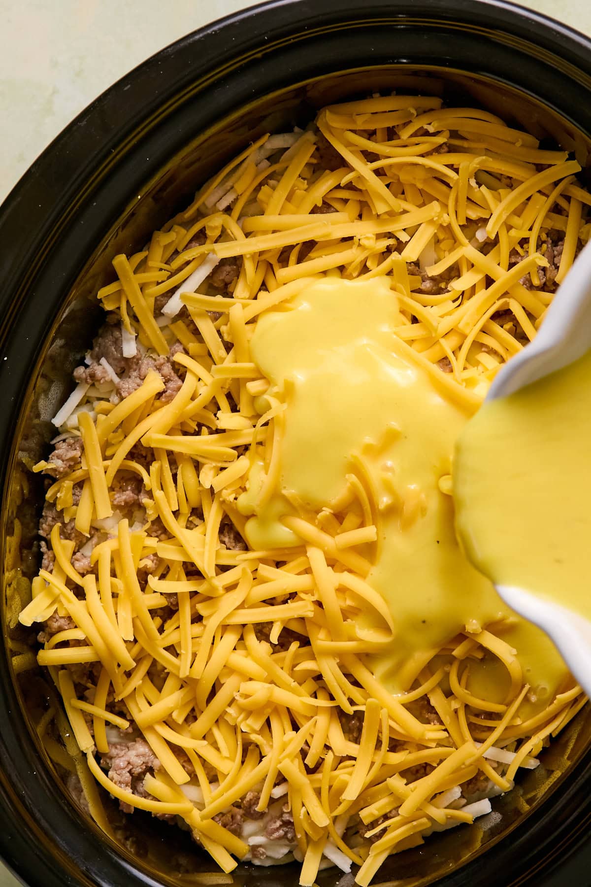 Egg mixture being poured over frozen hash browns, browned breakfast sausage, and cheese.
