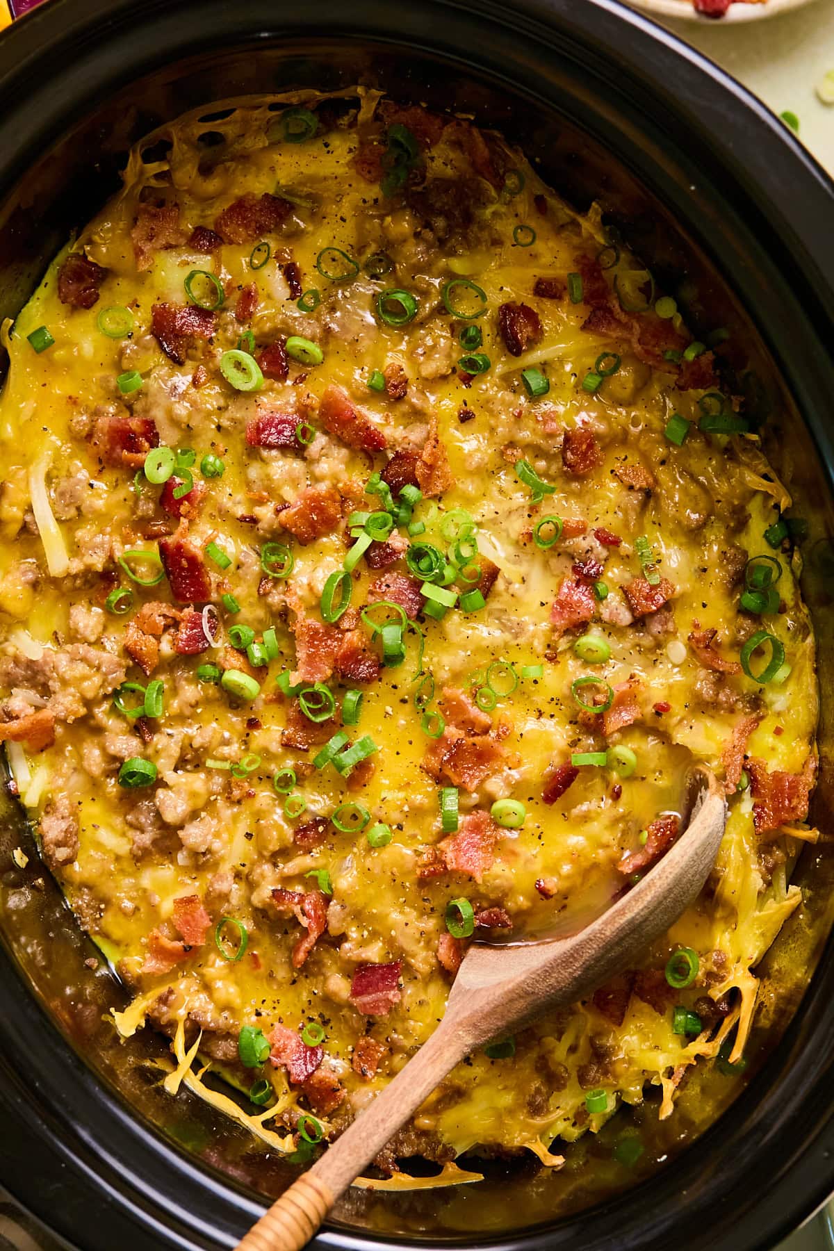 Close-up of breakfast casserole in a crockpot topped with crispy bacon and green onions. A wooden spoon is resting in the casserole.