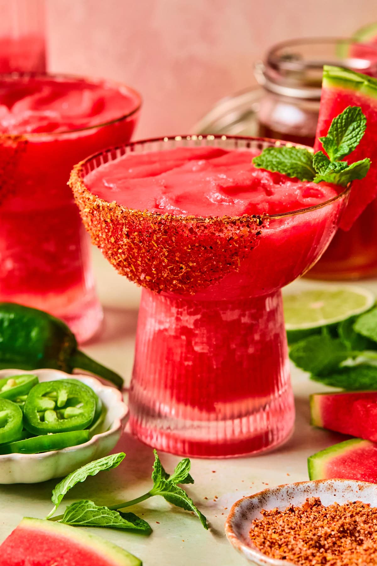 Two glasses of watermelon margaritas with a hot honey Tajín rim, garnished with fresh basil and watermelon slices, surrounded by fresh watermelon, basil, lime slices, and jalapeños.