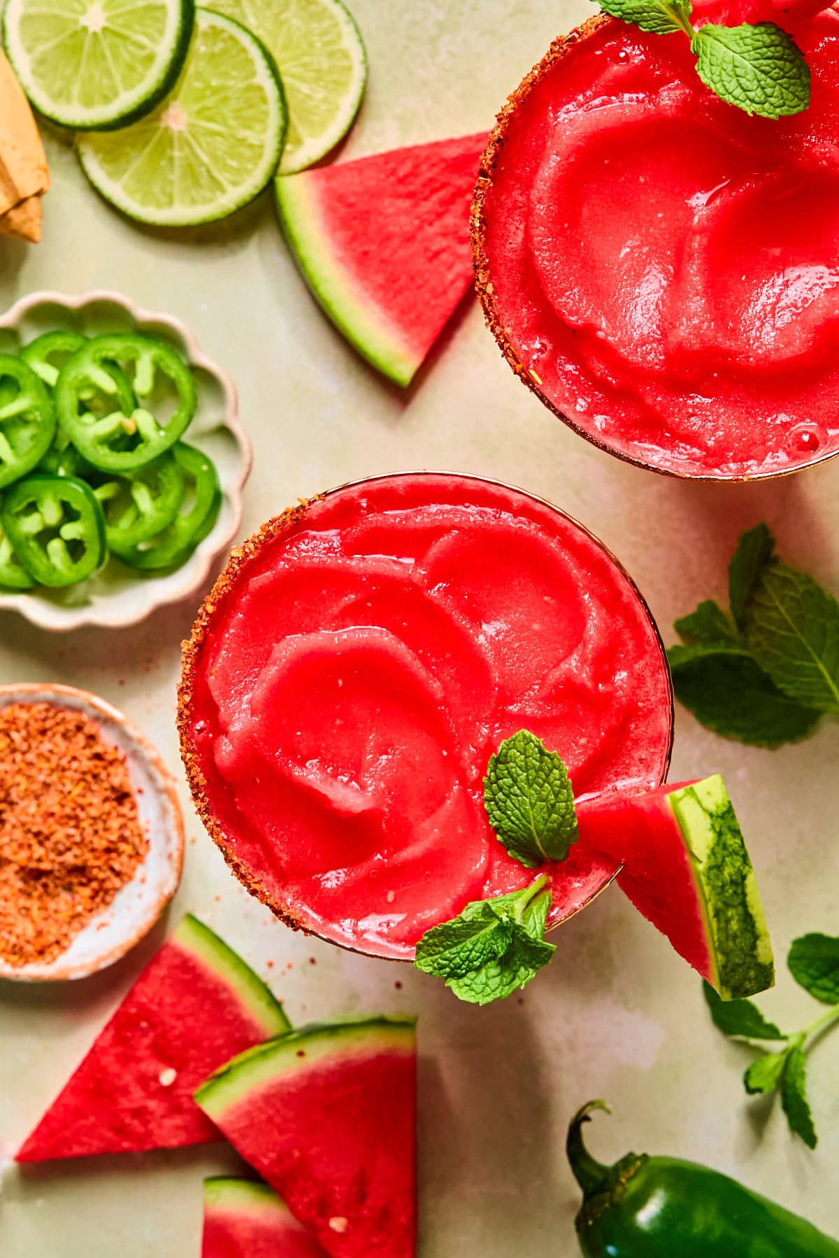 Overhead view of two watermelon margaritas with a hot honey Tajín rim, garnished with fresh basil and watermelon slices, surrounded by fresh watermelon, basil, lime slices, and jalapeños.