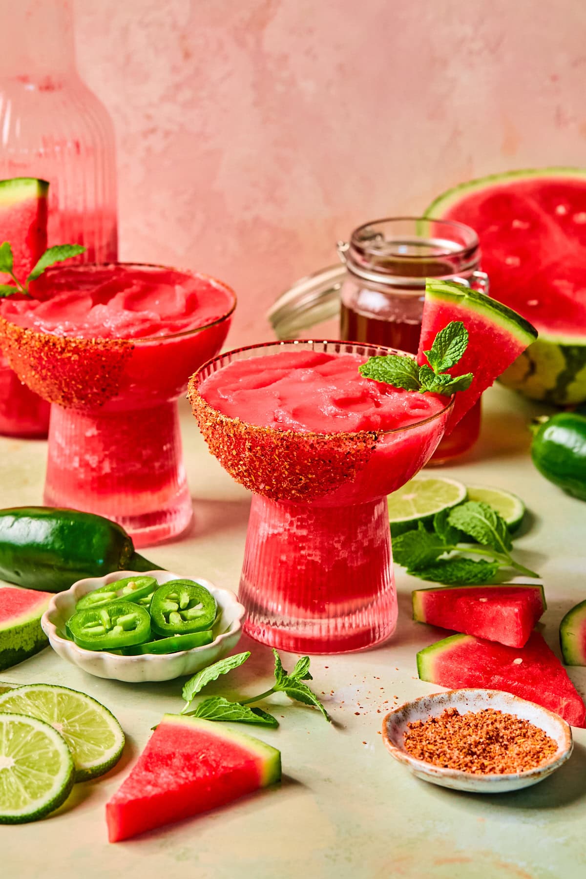 Two glasses of watermelon margaritas with a hot honey Tajín rim, garnished with fresh basil and watermelon slices, surrounded by fresh watermelon, basil, lime slices, and jalapeños.
